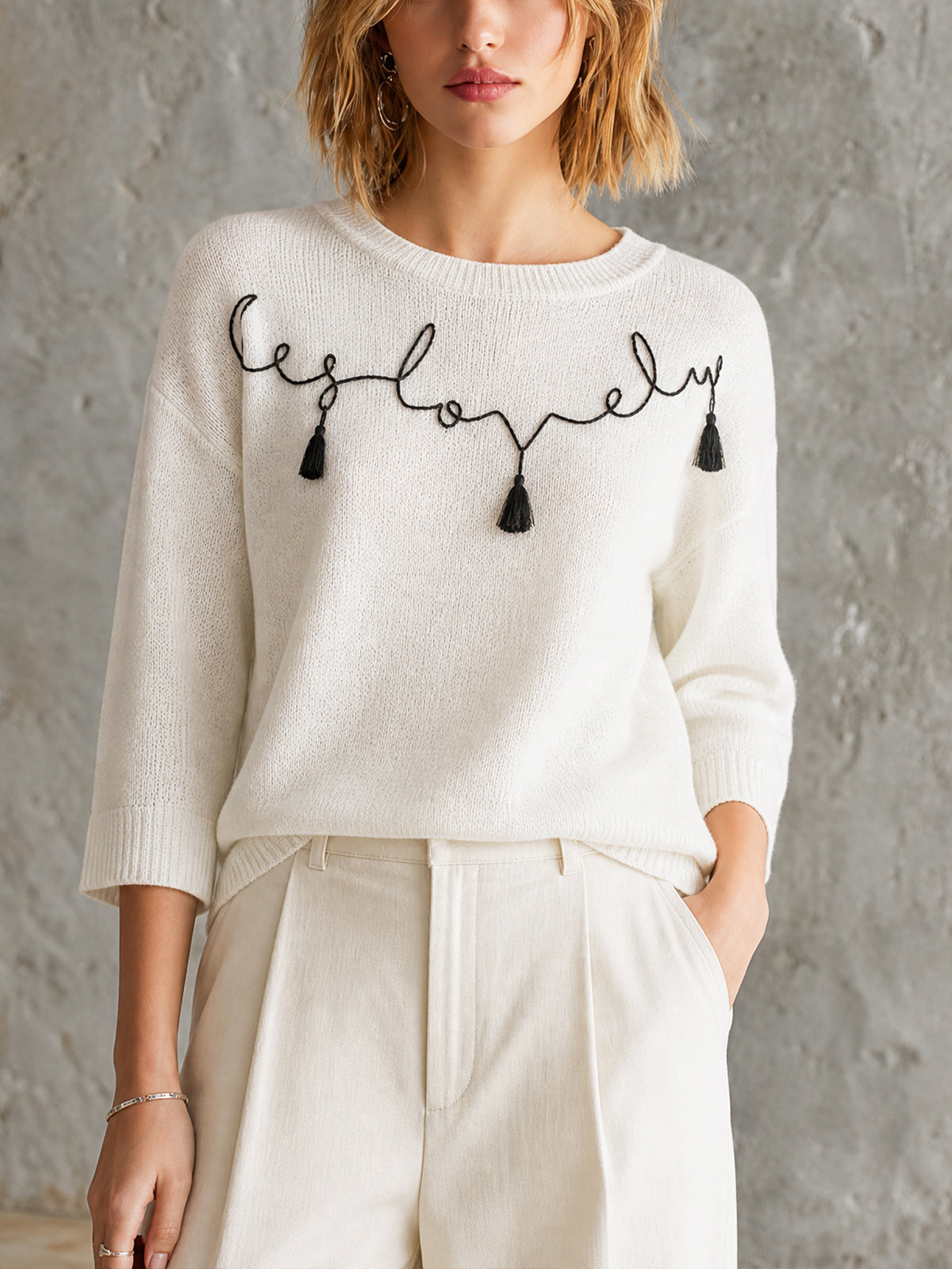 Pull écru motif manuscrit — vue portée de face, inscription brodée noire, pantalon blanc