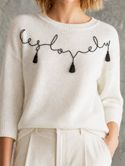 Pull écru motif manuscrit — gros plan de la broderie « be lovely » et des pampilles noires