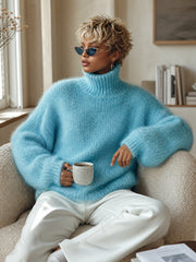 Pull texturé à col montant bleu ciel — vue portée assise dans un fauteuil blanc, mug à la main
