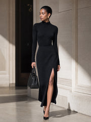 Robe longue noire fendue, portée devant une façade claire.