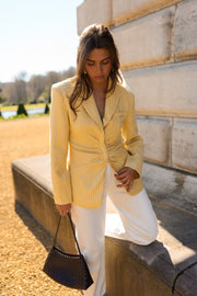 Blazer rayé cintré à épaulettes porté par une femme, vue de trois-quarts, pantalon blanc, sac noir, sur un muret