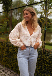 Blouse fleurie à décolleté dentelle portée par une femme, silhouette entière, pergola, mains dans les poches