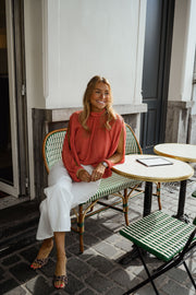 Blouse froncée à col montant terracotta portée par une femme, assise, à une table, pantalon blanc