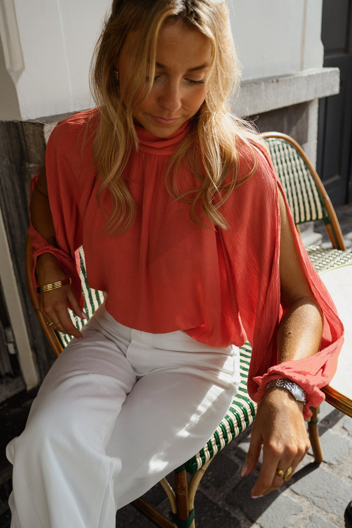 Blouse froncée à col montant terracotta portée par une femme, assise, sur une chaise de café, pantalon blanc