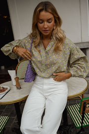 Blouse fleurie légèrement évasée portée par une femme, assise, à une table, sac violet