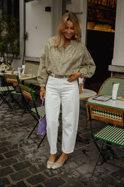 Blouse fleurie légèrement évasée portée par une femme, silhouette entière, sac violet, pantalon blanc, terrasse