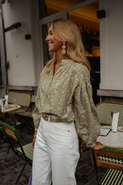 Blouse fleurie légèrement évasée portée par une femme, silhouette entière, pantalon blanc, terrasse