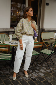 Blouse fleurie légèrement évasée portée par une femme, vue de trois-quarts, sac violet, pantalon blanc, terrasse