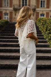 Blouse en dentelle à volants portée par une femme, vue de dos, escalier, pantalon blanc