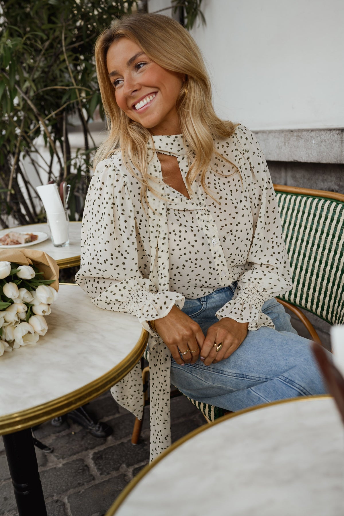 Blouse à pois col lavallière portée par une femme, assise, à une table, fleurs blanches, jean clair