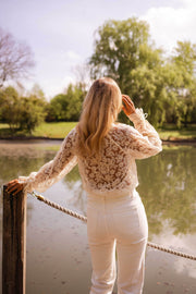Blouse transparente col V boutonnée écrue portée par une femme, vue de trois-quarts, ponton en bois, pantalon blanc