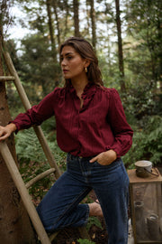 Blouse brodée à col volanté bordeaux portée par une femme, vue de trois-quarts, échelle en bois, mains dans les poches