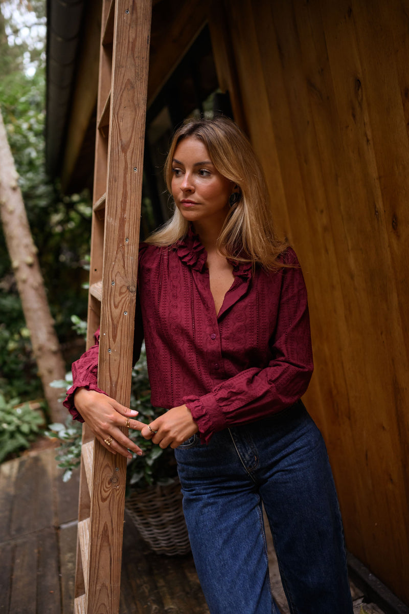 Blouse brodée à col volanté bordeaux portée par une femme, vue de face, poteau en bois, jean bleu