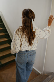 Blouse fleurie fluide à volants portée par une femme, vue de dos, main sur le mur, queue de cheval