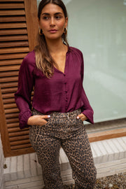 Blouse satinée col V bordeaux portée par une femme, vue de face, volets en bois, pantalon léopard