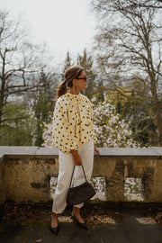 Blouse à pois dos ouvert portée par une femme, vue de trois-quarts, pantalon blanc, sac noir, devant un mur