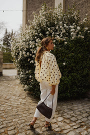 Blouse à pois dos ouvert portée par une femme, vue de trois-quarts, sac noir, devant un buisson fleuri
