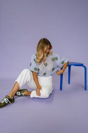 Blouse plissée col V portée par une femme, assise, sur un tabouret bleu, vue de côté