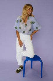 Blouse plissée col V portée par une femme, silhouette entière, pantalon blanc, pied sur un tabouret bleu