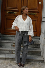 Blouse en dentelle col V portée par une femme, silhouette entière, jean gris, devant une porte en bois
