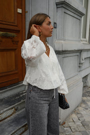 Blouse en dentelle col V portée par une femme, vue de trois-quarts, jean gris, porte en bois, main dans les cheveux