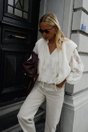 Blouse en dentelle col V portée par une femme, vue de trois-quarts, lunettes de soleil, pantalon clair, porte noire