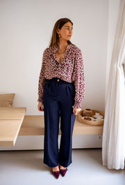 Blouse imprimée fluide portée par une femme, vue de face, banquette en bois, pantalon bleu foncé