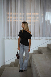 Blouse à épaule dénudée noire portée par une femme, vue de face, escalier, jean clair