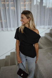 Blouse à épaule dénudée noire portée par une femme, vue de trois-quarts, rideaux blancs, sac noir