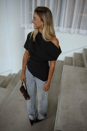 Blouse à épaule dénudée noire portée par une femme, vue de trois-quarts, marches grises, regard de côté