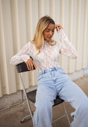 Blouse moulante en dentelle écrue portée par une femme, assise, mur beige, jean clair