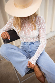 Blouse moulante en dentelle écrue portée par une femme, assise, chapeau beige, pochette noire