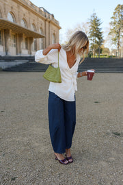 Blouse cache-cœur fluide portée par une femme, vue de trois-quarts, pantalon bleu marine, sac vert, gobelet à la main