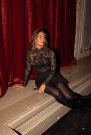 Blouse en dentelle transparente noire portée par une femme, assise, assise sur les marches, collants noirs