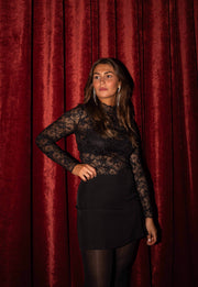 Blouse en dentelle transparente noire portée par une femme, silhouette entière, mini-jupe noire, rideau rouge