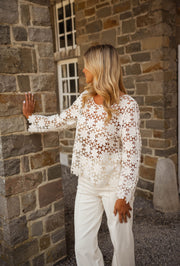 Blouse brodée à manches évasées écrue portée par une femme, vue de trois-quarts, mur en pierre, pantalon blanc
