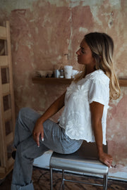 Blouse brodée à manches courtes écrue portée par une femme, assise, tabouret, mur patiné
