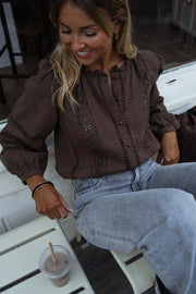 Blouse brodée à boutons chocolat portée par une femme, assise, banc blanc, jean clair