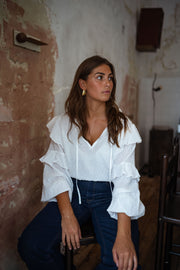 Blouse plumetis à volants écrue portée par une femme, vue de trois-quarts, mur patiné, jean foncé