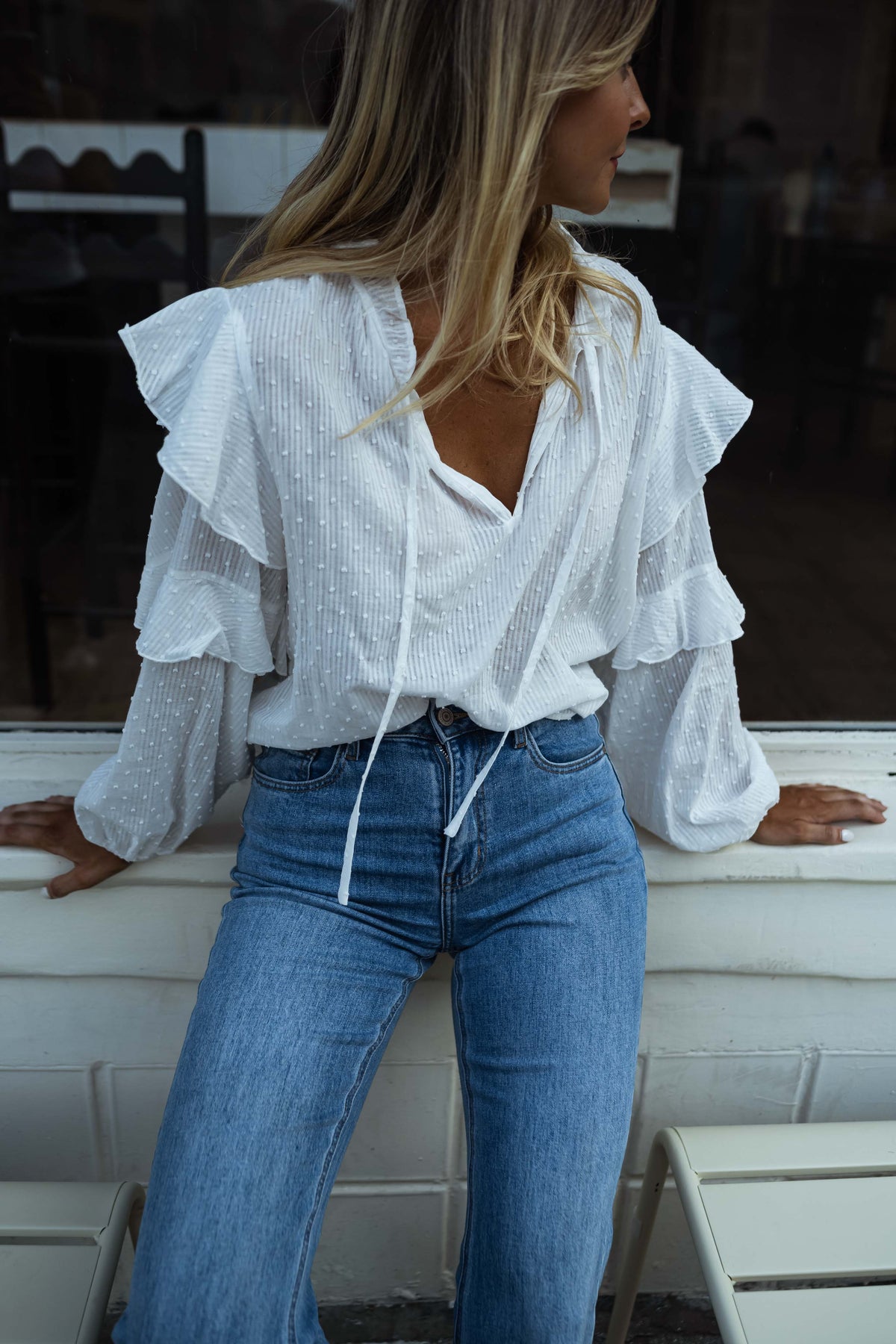 Blouse plumetis à volants écrue portée par une femme, vue de face, banc blanc, jean bleu