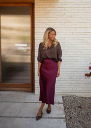 Chemise satinée fluide chocolat portée par une femme, silhouette entière, porte en bois, jupe