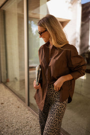 Chemise manches longues loose chocolat portée par une femme, vue de profil, baie vitrée, pantalon léopard