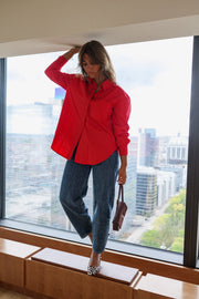 Chemise ample à manches longues rouge portée par une femme, silhouette entière, près d'une fenêtre