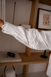 Chemise effet lin brodée blanche portée par une femme, vue rapprochée, bras tendu sur fond d'étagère
