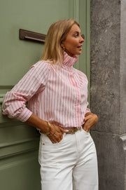Chemise rayée à col victorien portée par une femme, vue de trois-quarts, adossée à un mur, pantalon blanc