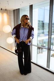 Chemise ample à carreaux portée par une femme, vue de face, pull bordeaux sur les épaules, baie vitrée