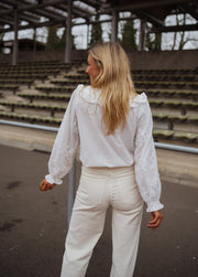 Chemise brodée col claudine blanche portée par une femme, vue de dos, gradins, pantalon blanc