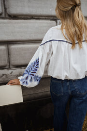 Chemise brodée à col montant portée par une femme, vue de dos, mur en pierre, broderie bleue