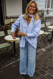 Chemise rayée à cravate portée par une femme, silhouette entière, jean large, sac marron, terrasse