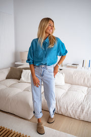 Chemise fluide effet satin bleue portée par une femme, vue de face, canapé blanc, pas de côté